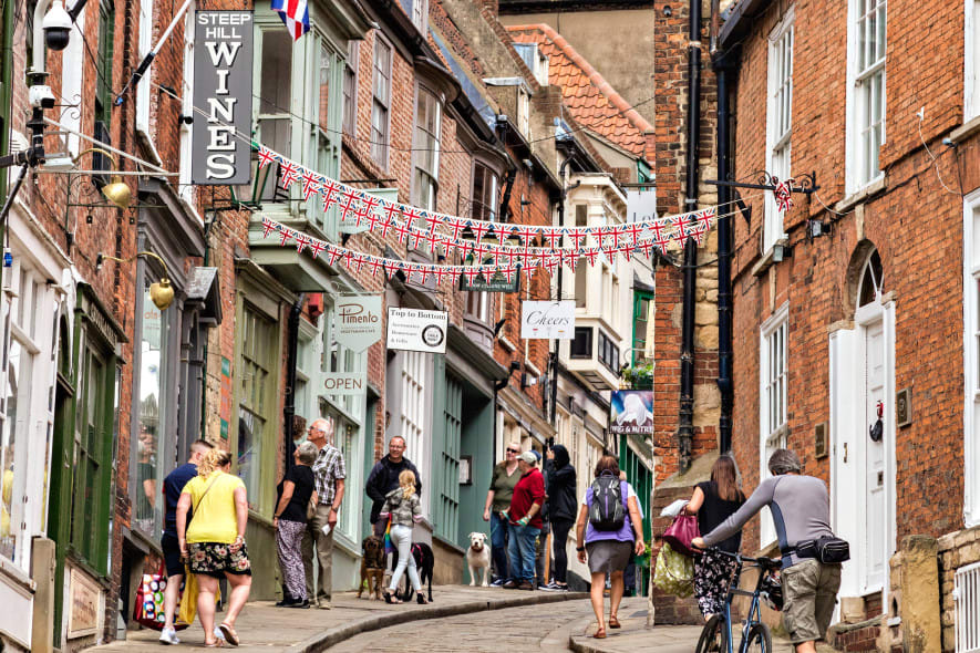 People stroll and shop along a steep cobbled street lined with historic brick buildings, Union Jack bunting, and quaint shopfronts.