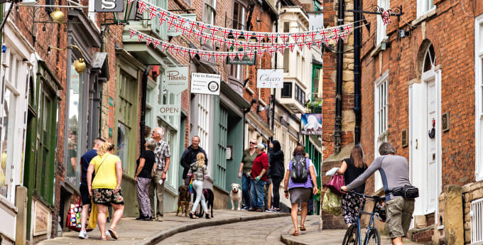 People stroll and shop along a steep cobbled street lined with historic brick buildings, Union Jack bunting, and quaint shopfronts.