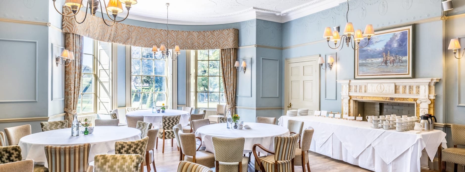 Light-filled event room with round tables, chandelier lighting, and an ornate fireplace set for refreshments.