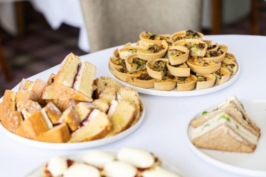 Afternoon tea table with Victoria sponge slices, mini quiches, sandwiches, and scones with cream and jam.