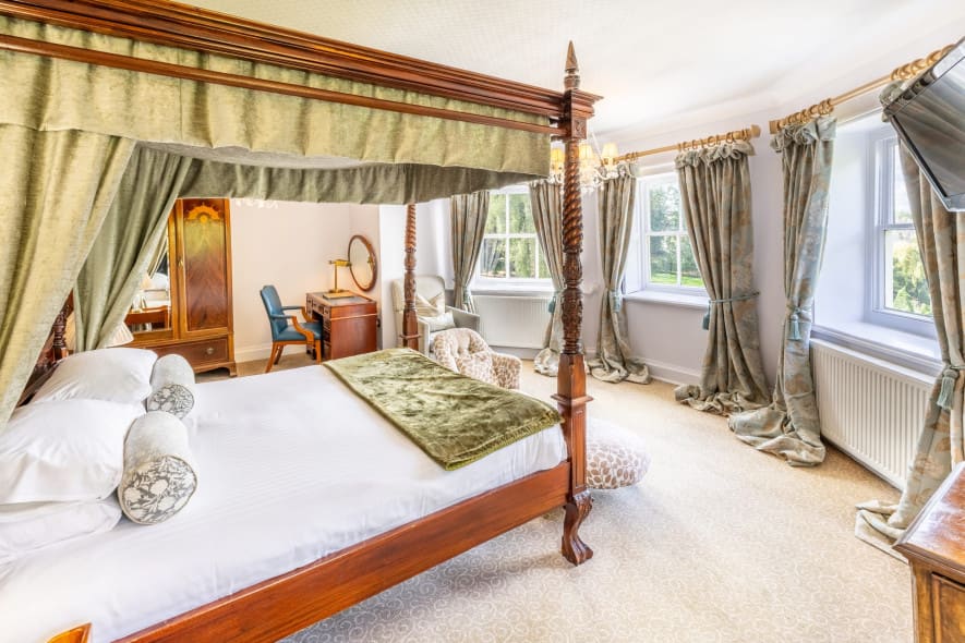 Bright, spacious hotel bedroom with a four-poster bed, antique wooden furniture, and large bay windows dressed with patterned curtains.