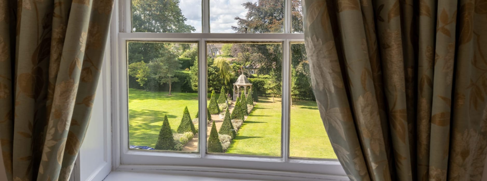 View through a sash window framed by elegant floral curtains, overlooking a formal garden with a row of conical topiary trees leading to a gazebo on the lawn.