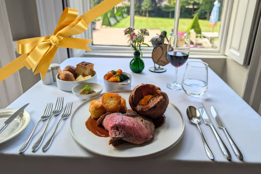 Elegant Sunday roast with beef, roast potatoes and Yorkshire pudding served at a white-tablecloth restaurant table with a gold gift ribbon overlay.