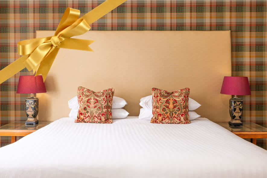 Plush double bed with patterned cushions, bedside lamps and tartan wallpaper, overlaid with a gold gift ribbon.