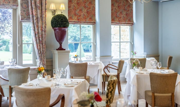 Corner of a restaurant with floral blinds, topiary decor, and elegant dining tables set for service near windows.