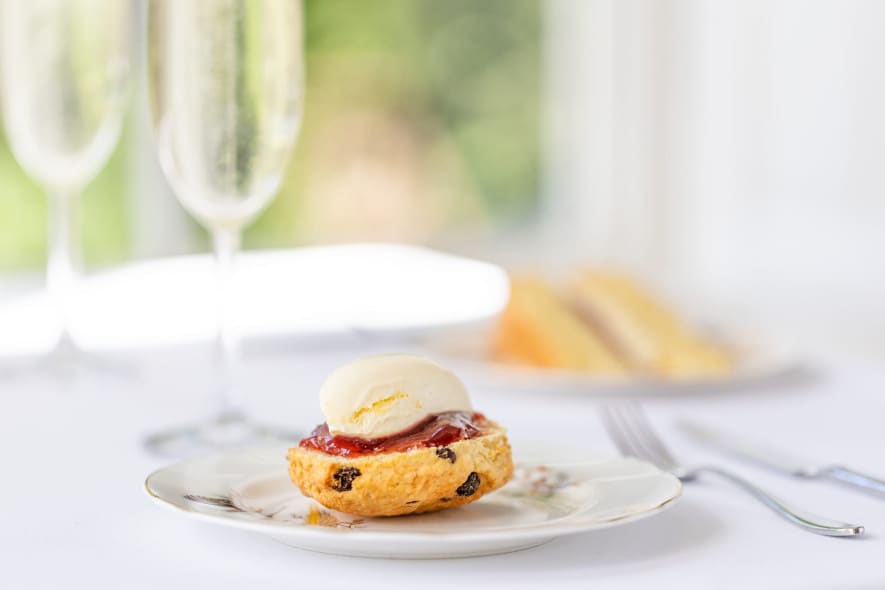 A scone topped with strawberry jam and clotted cream on a floral plate, with champagne flutes in background.