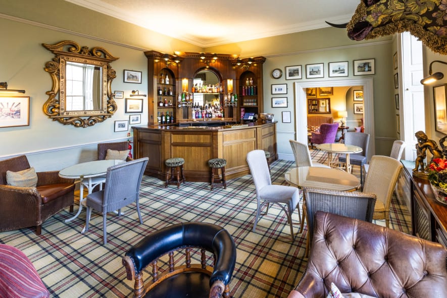 Traditional hotel bar with wood paneling, tartan carpet, mirror, armchairs, and shelves stocked with spirits.