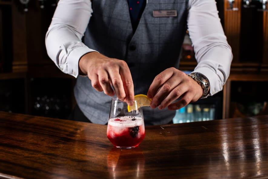 Close-up of bartender garnishing red cocktail with lemon wedge, hands mid-motion over wood bar counter.
