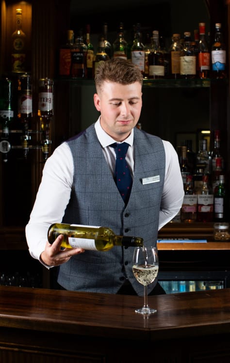 Bartender pours white wine into glass at hotel bar, surrounded by spirits, champagne bucket, and soft warm light.
