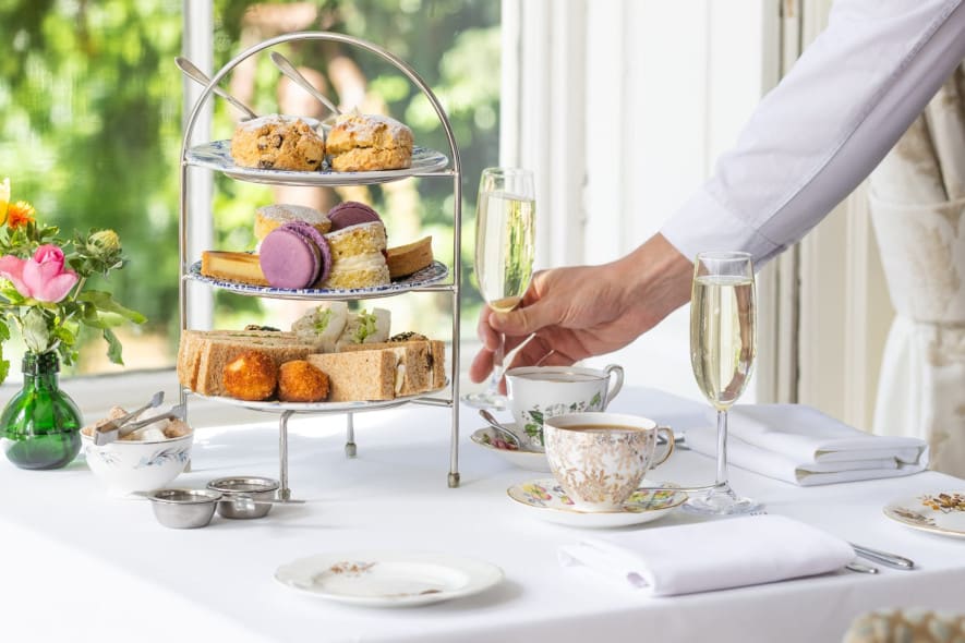 A server in a white shirt places a champagne flute on a table set for afternoon tea with tiered treats, fine china, and fresh flowers near a sunlit window.
