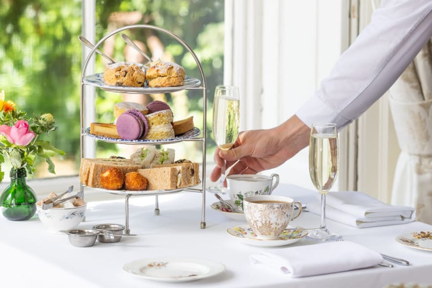 A server in a white shirt places a champagne flute on a table set for afternoon tea with tiered treats, fine china, and fresh flowers near a sunlit window.