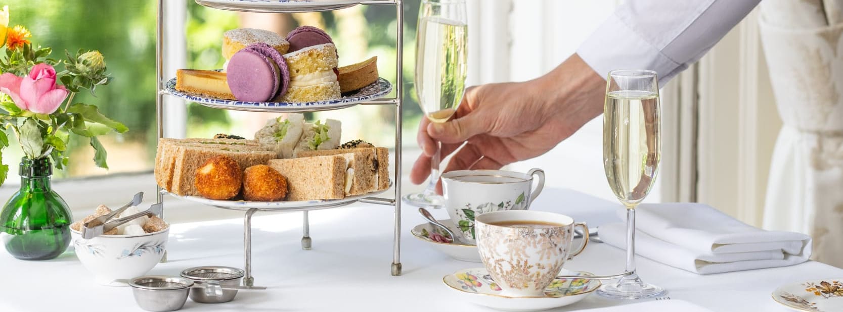 A server in a white shirt places a champagne flute on a table set for afternoon tea with tiered treats, fine china, and fresh flowers near a sunlit window.