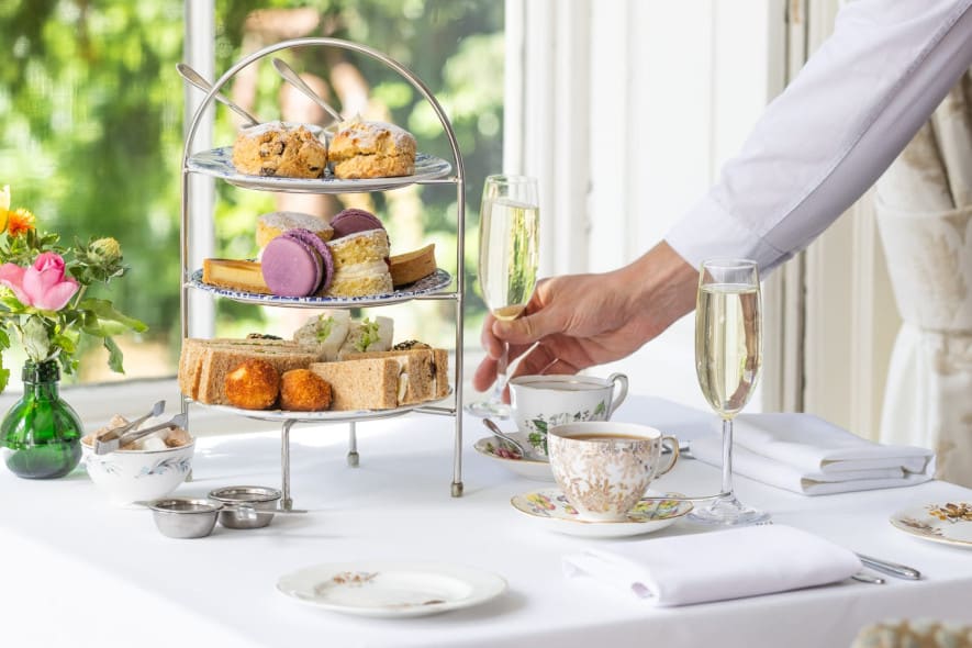 A server in a white shirt places a champagne flute on a table set for afternoon tea with tiered treats, fine china, and fresh flowers near a sunlit window.