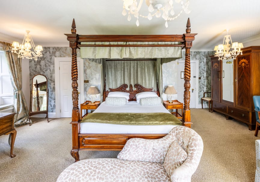 Elegant bedroom with a carved four-poster bed, soft green drapes, vintage furniture, and multiple windows letting in natural light.