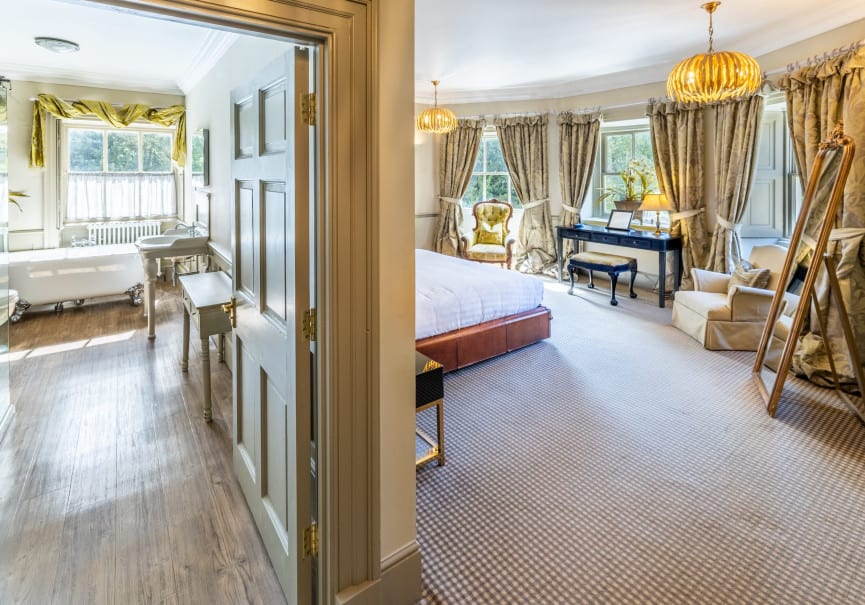 View from hallway into a bright suite with a classic bathroom on one side and a window-lit bedroom with elegant furnishings on the other.