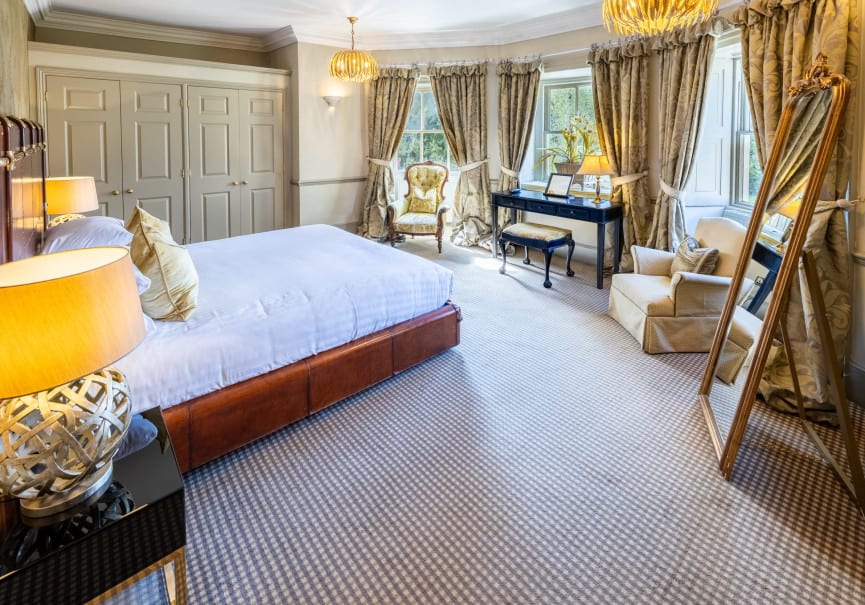 Bright, traditional-style bedroom with a large bed, plush carpet, gold-toned accents, and a wall of windows dressed in patterned curtains.