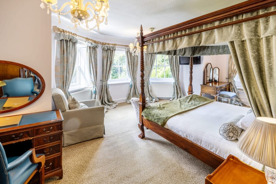 Corner view of a stately bedroom with a canopy bed, chandelier, writing desk, and tall windows draped in floral curtains.