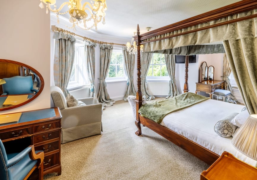 Corner view of a stately bedroom with a canopy bed, chandelier, writing desk, and tall windows draped in floral curtains.