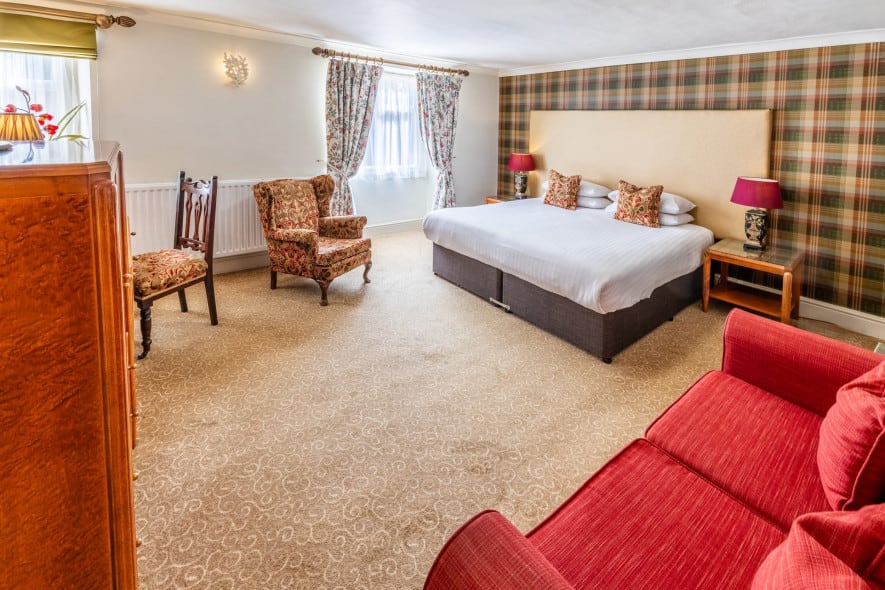 Spacious traditional hotel bedroom with super king bed, red sofa, floral armchairs and tartan feature wall, lit by natural light from two windows.