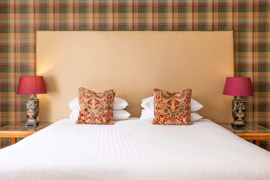 Super king hotel bed with tall cream headboard, tartan feature wall, patterned cushions and matching bedside lamps with rich red shades.