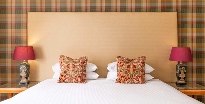 Super king hotel bed with tall cream headboard, tartan feature wall, patterned cushions and matching bedside lamps with rich red shades.