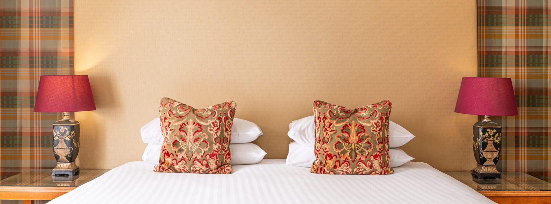 Super king hotel bed with tall cream headboard, tartan feature wall, patterned cushions and matching bedside lamps with rich red shades.