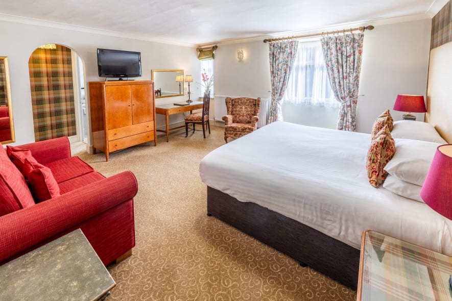 Traditional hotel bedroom with super king bed, tartan feature wall and bedside lamp, viewed from across the room near a wooden wardrobe.