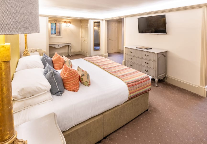 Spacious country-style bedroom with super king bed, colourful cushions, grey dresser, wall-mounted TV and soft lighting.