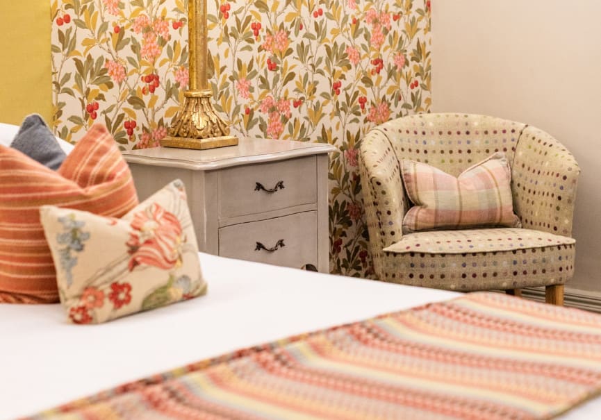 Floral feature wall with gold lamp, grey bedside cabinet and patterned tub chair beside a super king hotel bed.