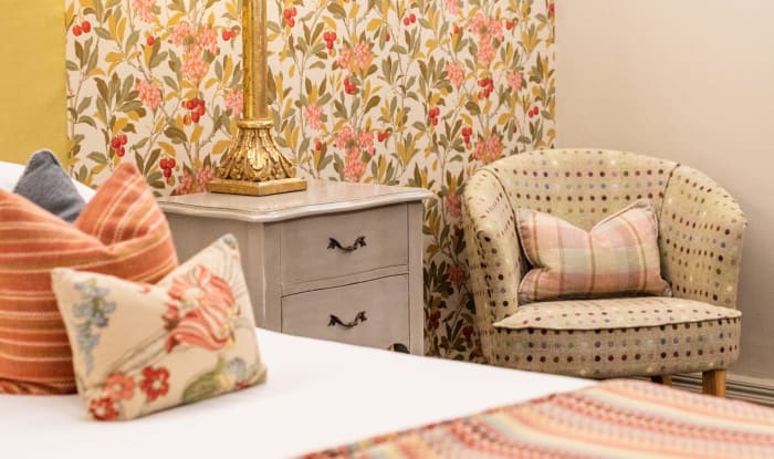 Floral feature wall with gold lamp, grey bedside cabinet and patterned tub chair beside a super king hotel bed.
