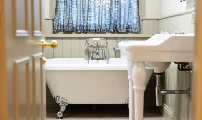 Roll-top bathtub with chrome feet and mixer taps, viewed from doorway beside a white pedestal sink and striped window curtain.