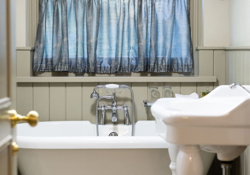 Cosy bathroom with roll-top bathtub, pedestal sink and window dressed with checked and striped curtains.
