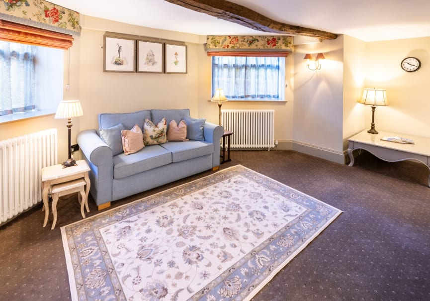 Bright sitting area with blue sofa, patterned rug, table lamps and exposed wooden beam above two windows.