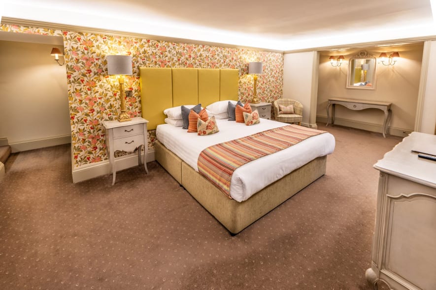 Country-house bedroom with super king bed, yellow headboard, floral wallpaper, bedside lamps and dressing table.