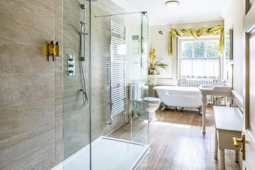 Alt text: Bright bathroom in the Superior Ingilby Room at Washingborough Hall with walk-in shower, roll-top bath, pedestal sink and window with green drapes.