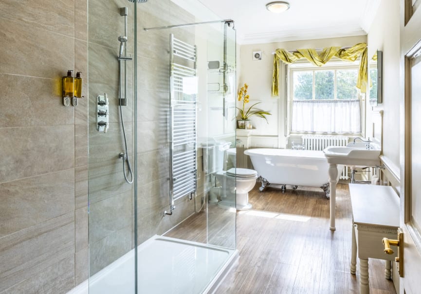 Alt text: Bright bathroom in the Superior Ingilby Room at Washingborough Hall with walk-in shower, roll-top bath, pedestal sink and window with green drapes.