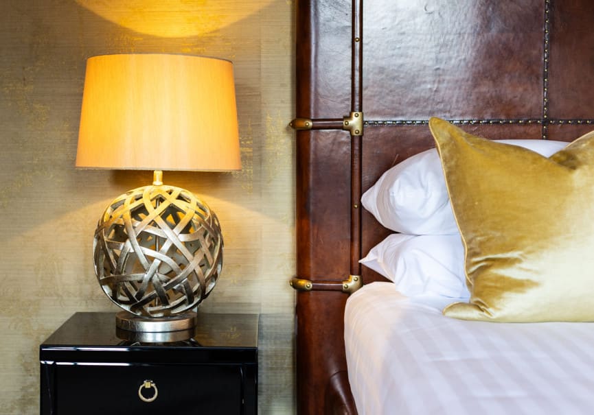 Bedside lamp and leather headboard in the Superior Ingilby Room at Washingborough Hall, with gold velvet cushion on crisp white bedding.