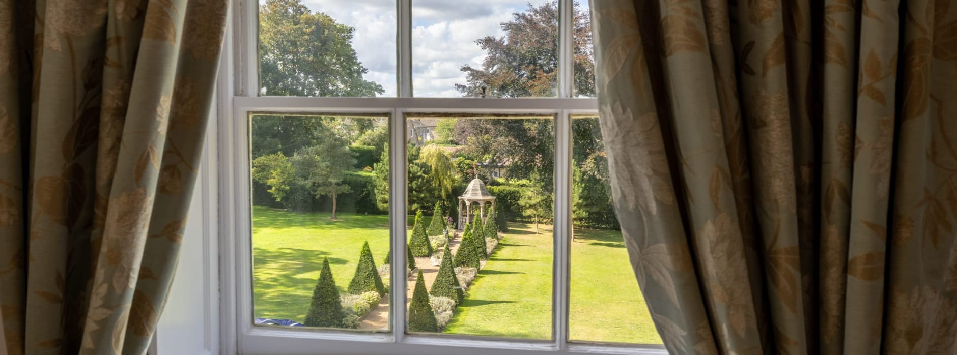 Garden view from the Superior Ingilby Room at Washingborough Hall, framed by floral curtains, overlooking lawns, topiary trees and a gazebo.