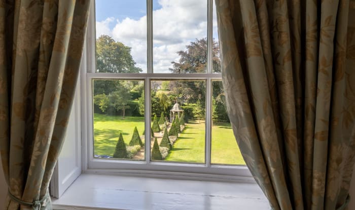 Garden view from the Superior Ingilby Room at Washingborough Hall, framed by floral curtains, overlooking lawns, topiary trees and a gazebo.