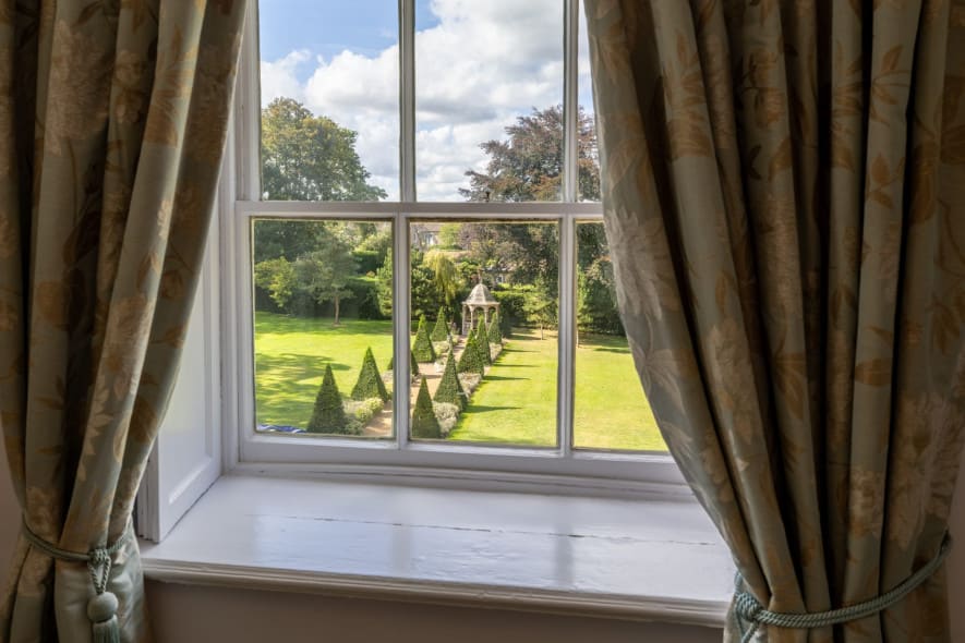 Garden view from the Superior Ingilby Room at Washingborough Hall, framed by floral curtains, overlooking lawns, topiary trees and a gazebo.
