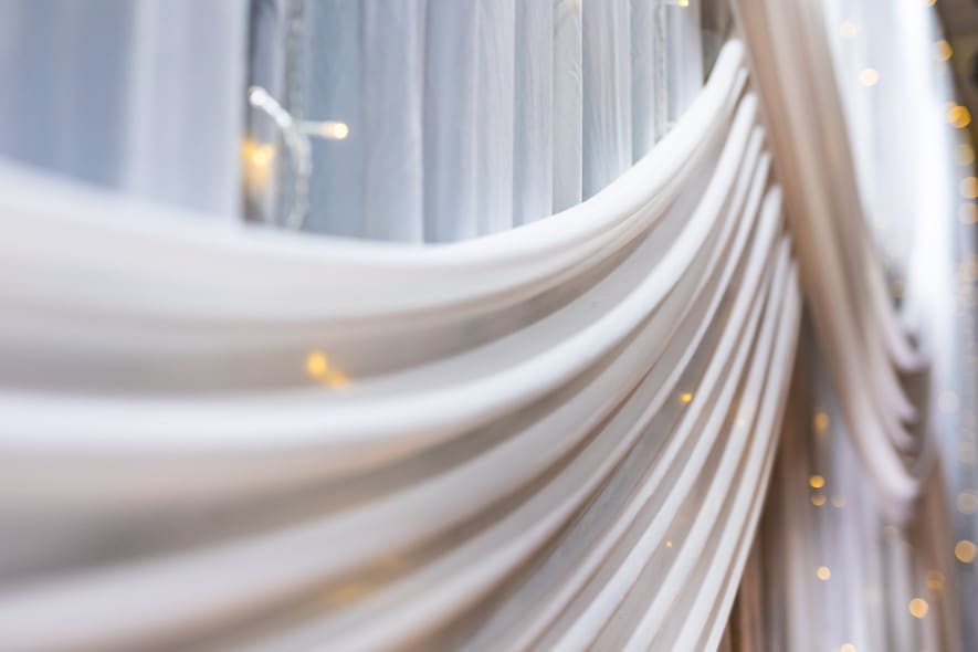 Close-up of elegant white draped fabric with soft folds and warm fairy lights, creating a dreamy and romantic backdrop, likely for a wedding or event.