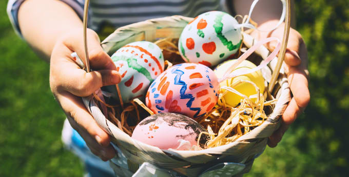 An image of a basket being held in a pair hands, which is full of colourfully painted easter eggs with straw underneath.