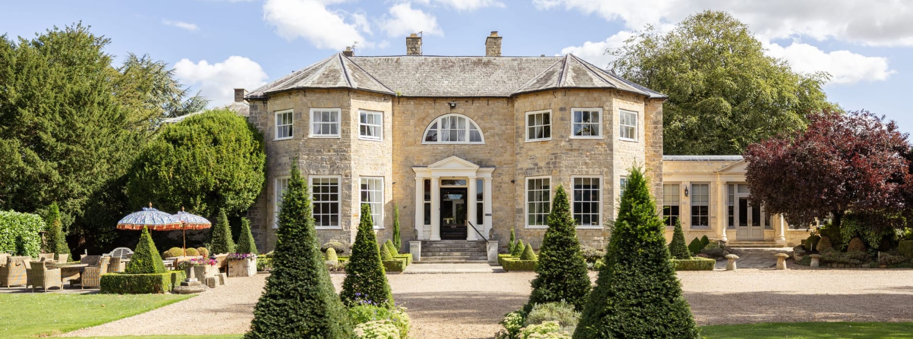 Washingborough Hall Hotel’s stone façade with symmetrical wings, formal garden, gravel path, and central fountain on a bright, clear day.