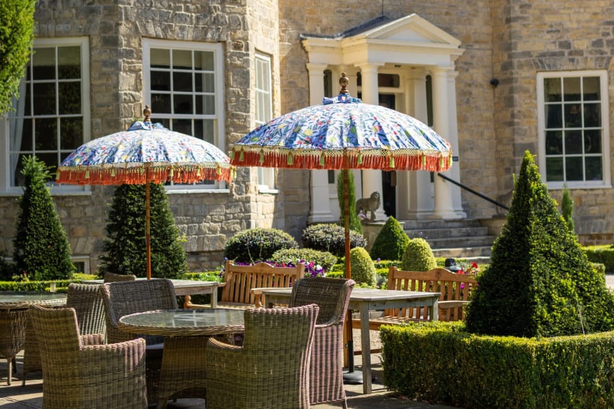 Washingborough Hall Hotel’s stone façade with patio seating, wicker furniture, colourful parasols, and manicured topiary on a bright summer day.