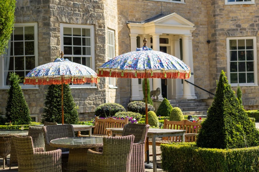 Washingborough Hall Hotel’s stone façade with patio seating, wicker furniture, colourful parasols, and manicured topiary on a bright summer day.