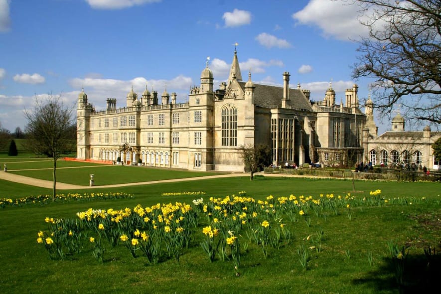 Burghley House, an ornate Elizabethan estate with towers and chimneys, rises above sweeping lawns beneath a bright blue sky with soft scattered clouds.