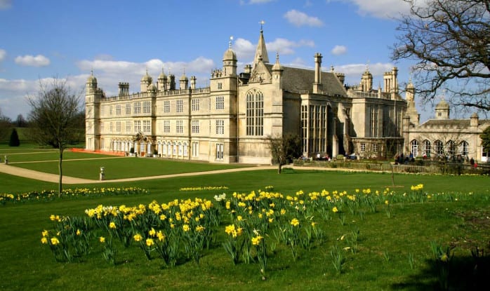 Burghley House, an ornate Elizabethan estate with towers and chimneys, rises above sweeping lawns beneath a bright blue sky with soft scattered clouds.