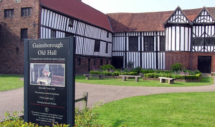 Gainsborough Old Hall, a medieval timber‑framed manor with brick and black‑and‑white façades, stands beside gardens and benches under a bright sky.