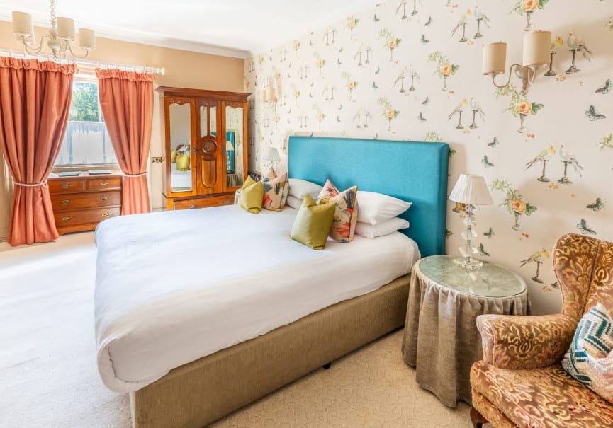 A luxurious room, featuring a large bed with a blue headboard, elegant wall paper and deep red curtains.