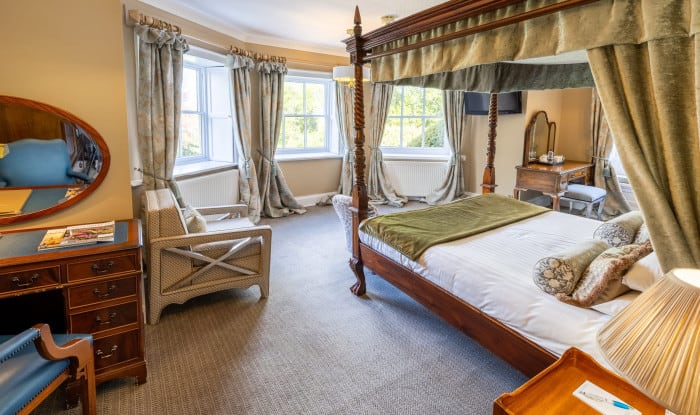 Elegant bedroom with four‑poster bed, bay windows, soft curtains, and antique furniture in warm beige tones.